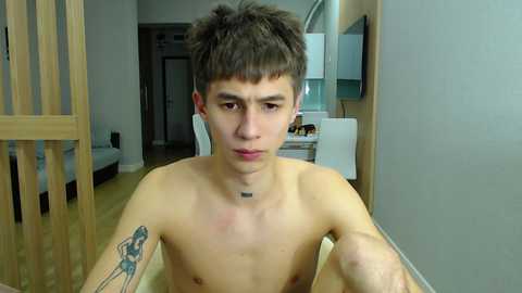 Media: Video of a shirtless young man with short, dark hair and a tattoo on his right arm, sitting in a modern apartment with light wooden floors, a kitchen in the background.