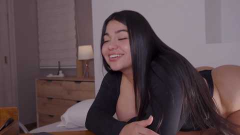 Media: Video of a smiling, voluptuous Latina woman with long black hair, lying on a bed in a simple bedroom. She wears a black top and black panties.