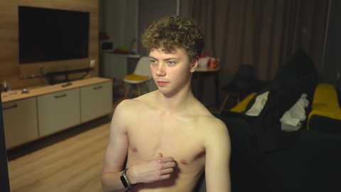 Media: Video of a shirtless, slender, young man with curly brown hair, standing indoors, wearing a smartwatch. Background includes a flat-screen TV, wooden cabinets, and a messy living room.