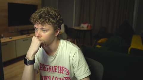 Media: Video of a young man with curly hair, wearing a white t-shirt with Greek text, sitting contemplatively in a dimly lit living room with a TV, a table, and a dark sofa.