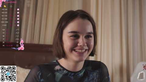 Media: Video of a smiling young woman with straight brown hair, wearing a black floral-patterned blouse. Background shows a beige curtain and a bed with white sheets.