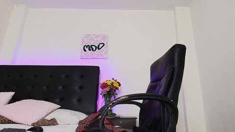 Media: Video of a modern bedroom with a black leather chair, a black upholstered headboard, and a floral arrangement on a wooden nightstand; a framed wall art piece hangs above.