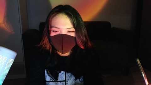 Media: Video of an Asian woman with shoulder-length, dark hair, wearing a black mask, black top, and a white graphic shirt, sitting in a dimly lit room with warm, colorful lighting.