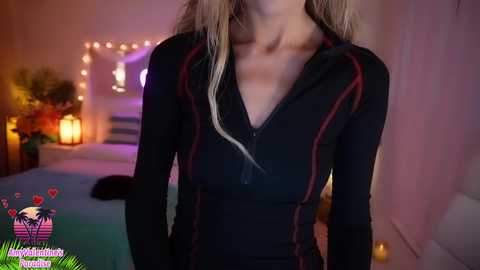 Media: Video of a slim, blonde woman in a black, long-sleeve, zip-up top, standing in a softly lit, cozy bedroom with string lights, a bed, and a floral arrangement in the background.