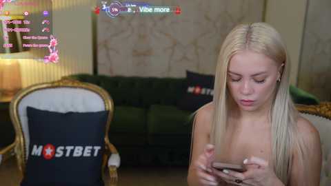 Media: Video of a blonde woman in a dark room, looking at a smartphone, wearing a black pillow with \"FSTBET\" text.