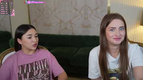 Media: Video of two young women with light skin, one in a pink \"JIMI HENDRIX\" T-shirt and the other in a white graphic tee, sitting in a cozy living room with green couch, floral wallpaper, and a lamp.