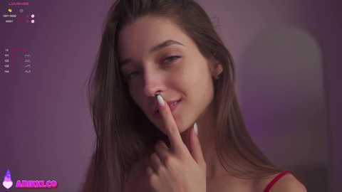 Media: Video of a young, fair-skinned woman with long brown hair, smiling with a finger to her lips. Background is a soft purple hue.