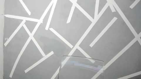 Media: Video of a minimalist, abstract wall with white, angular, overlapping lines on a light gray background, featuring a clear glass object at the bottom center.