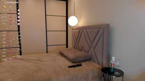 Media: Video of a modern, minimalistic bedroom with beige bedding, a padded headboard, and a spherical pendant light. A small, round side table holds a black remote and a glass.