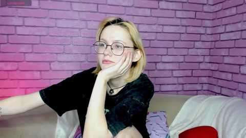 Media: Video of a young woman with blonde hair, glasses, and a black t-shirt, resting her head on her hand against a pink brick wall background.