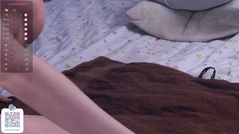 Media: Video of a person's hand holding a smartphone displaying a \"Loudness\" app, with a brown blanket partially covering them. The background shows a white bed with floral sheets and a beige pillow.