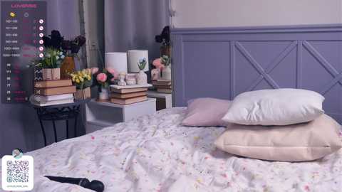 Media: A video of a neatly made bed with pastel-colored pillows, adorned with floral bedding, against a purple-paneled wall. A small table with books and flowers is in the background.