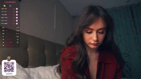 Media: A video of a young woman with long, wavy black hair and fair skin, wearing a red top, sitting on a bed with a gray headboard. The room has a dim, moody atmosphere with muted tones.