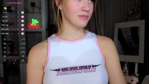 Media: Video of a young woman with light skin, brown hair, and a slim figure wearing a white tank top with pink trim, with a printed message across her chest, in a dimly lit room with a green neon sign and various objects in the background.