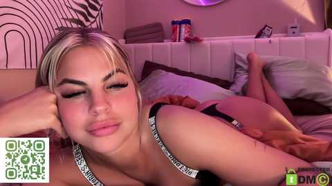 Media: Video of a blonde woman with large breasts and tanned skin lying on a bed, wearing a black bra. She has a tattoo on her left arm. The background features a white headboard and zebra-patterned wall art.