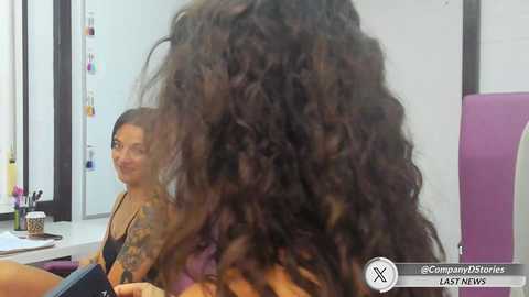 Media: Video of a woman with long, voluminous curly hair, sitting in a chair with a mirror behind her, smiling at her reflection. She wears a dark top and has tattoos on her arm.