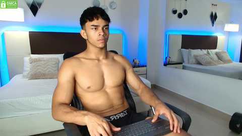 Media: Video of a shirtless, muscular, young Hispanic man with short dark hair, wearing black underwear, seated in front of a computer in a modern bedroom with white bedding, blue LED lighting, and a large mirror.