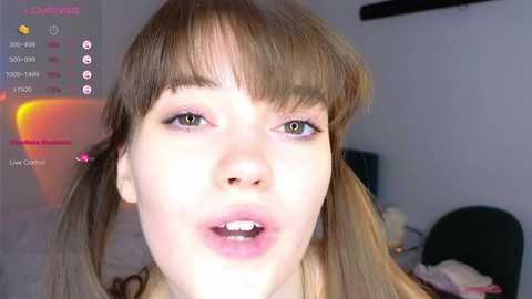 Media: Video of a young woman with light skin, straight light brown hair, and bangs. She has a surprised expression with slightly open mouth. The background shows a dimly lit room with a bed and a chair.