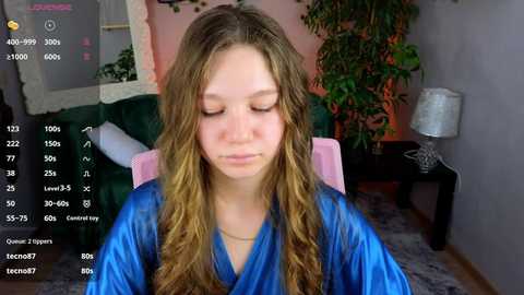 Media: Video of a young woman with long, wavy blonde hair, wearing a blue satin robe, sitting on a pink chair in a cozy room with a green bed, potted plants, and a lamp.