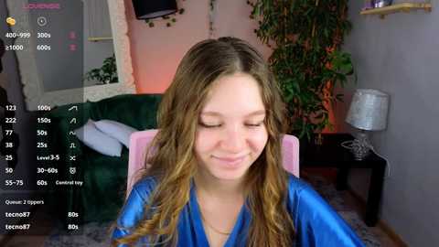 Media: Video of a young woman with long, wavy blonde hair, wearing a blue satin robe, smiling in a cozy, dimly-lit room with a green sofa, a lamp, and a plant.