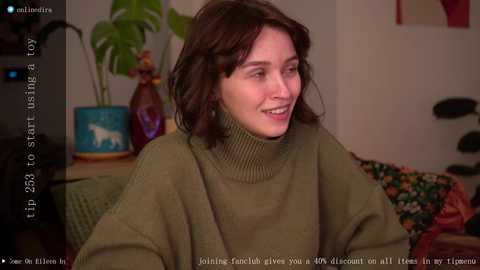 Media: A video of a woman with shoulder-length brown hair, smiling, wearing a green turtleneck sweater, sitting on a couch in a cozy living room with potted plants and a colorful blanket.