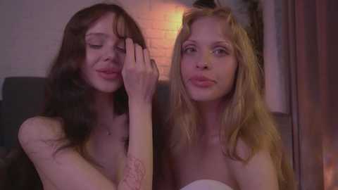 Media: Video of two young women with fair skin, one with long brown hair and the other with long blonde hair, both topless, smiling playfully, in a dimly lit room with brick walls and a warm, orange light.