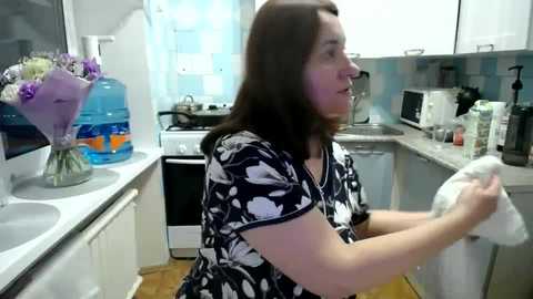 Media: Video of a middle-aged woman with long brown hair, wearing a navy floral dress, drying dishes in a small, bright kitchen with white cabinets, blue tiles, and a glass vase of purple flowers on the counter.