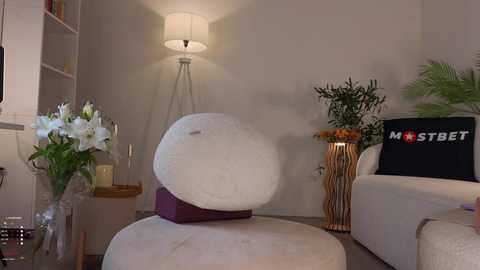 Media: Video of a modern living room with a white, cushioned, round chair, a tall floor lamp, a vase of white lilies, a black \"MSTBET\" pillow, and a wooden vase with greenery.