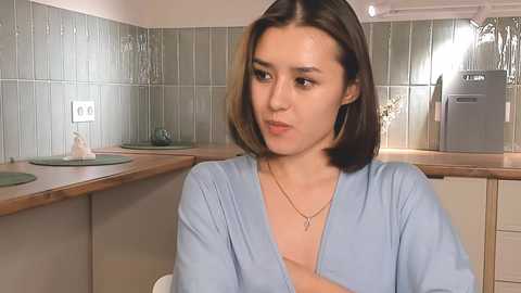 Media: A video of a young woman with shoulder-length brown hair, wearing a light blue V-neck shirt, sitting in a modern kitchen with tiled backsplash and white cabinets.