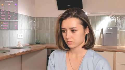 Media: Video of a young woman with short, straight brown hair, wearing a light blue robe, sitting in a modern kitchen with tiled backsplash and appliances.