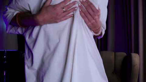 Media: Video of a person wearing a white robe, hands clasped over chest, against a purple-hued background with dark curtains and a green sofa.