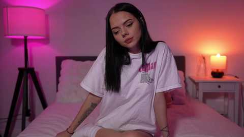 Media: Video of a young woman with long black hair, wearing a white t-shirt and sitting on a bed in a dimly lit bedroom with pink and orange lighting.