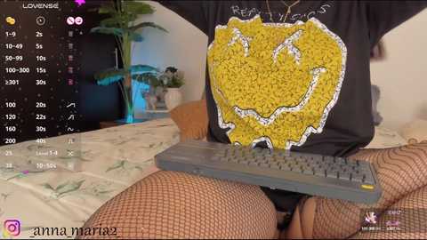 Media: Video of a person with a black T-shirt featuring a yellow map of the United States and black fishnet stockings, sitting on a bed with a green comforter. A gray keyboard is placed on their lap.