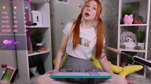 Media: Video of a young, pale-skinned woman with long red pigtails, wearing a white t-shirt with a cartoon graphic and a checkered skirt, playing a keyboard in a colorful, cluttered room.