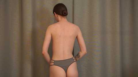 Media: A video of a shirtless woman with fair skin and a small tattoo on her right wrist, wearing a gray thong, standing with hands on hips against a gray curtain background.