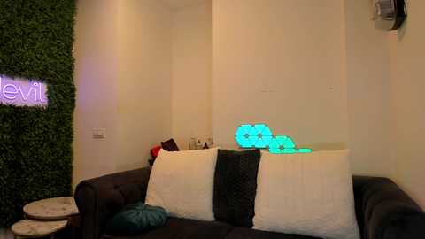 Media: Video of a cozy living room with a dark brown leather sofa adorned with white and black textured pillows, a vibrant green faux plant wall, a neon \"evil\" sign, and a glowing blue LED light on the wall.