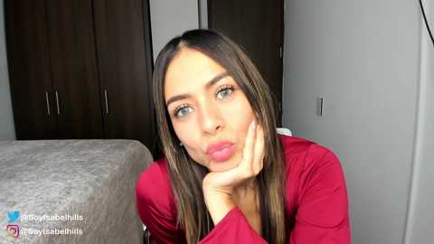 Media: A video of a young woman with long, straight brown hair, wearing a red top, resting her chin on her hand, looking contemplative. Background includes a bed and dark wooden wardrobe.