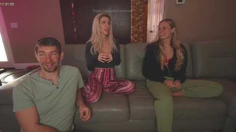 Media: Video of three people on a gray couch in a living room; two women with long blonde hair in casual attire, one man with short dark hair in a green shirt.