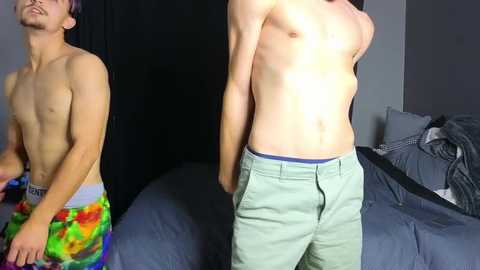 Media: Video of two shirtless teenage boys, one wearing vibrant floral shorts, standing in a dimly lit bedroom.