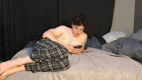 Media: Video of a shirtless, chubby man with short dark hair in black and white plaid pajama pants, lying on a bed with gray bedding, looking at a smartphone.