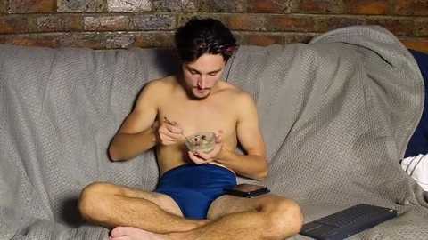 Media: Video of a shirtless man with a beard, wearing blue shorts, holding a small snake on his chest, sitting on a gray couch with a brick wall background.