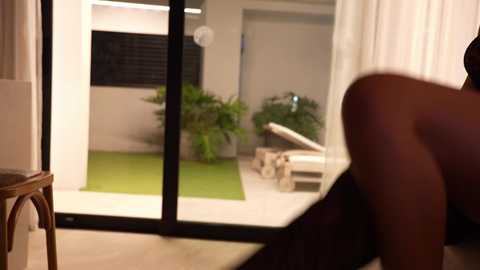 Media: Video of a bare-chested, dark-skinned man in white towel, standing near a large window overlooking a modern, minimalist garden with a green lawn, potted plants, and wooden furniture.