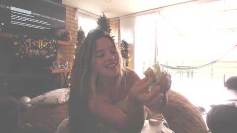 Media: Video of a joyful, topless woman with long brown hair, holding a banana, in a cozy, sunlit living room with a hammock, brick wall, and colorful TV stand.