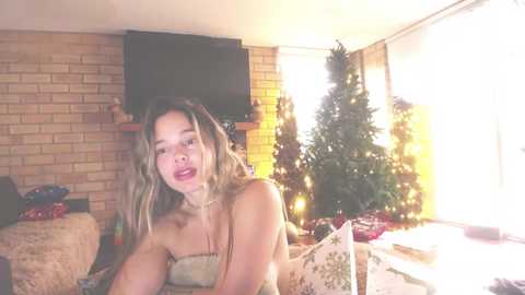 Media: A candid video of a young woman with long blonde hair, wearing a strapless top, sitting on a bed near a lit Christmas tree in a warmly lit, cozy room.