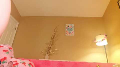 Media: Video of a cozy, warm-toned bedroom with a yellow wall, a white door, a floral bedspread, a decorative branch, a colorful poster, and a lamp with a pink shade.