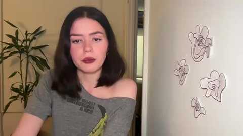 Media: A video of a young woman with straight, shoulder-length black hair, wearing a gray off-the-shoulder shirt, standing in a room with a white wall adorned with simple black flower doodles.