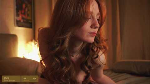 Media: Video of a young Caucasian woman with long, wavy red hair, fair skin, and large breasts, sitting on a bed in a dimly lit bedroom with warm, ambient lighting.