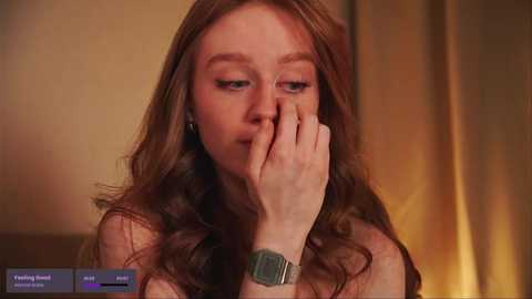 Media: Video of a fair-skinned, red-haired woman with long, wavy hair, wearing a silver wristwatch, thoughtfully covering her mouth with her right hand, in a dimly lit room with golden curtains in the background.