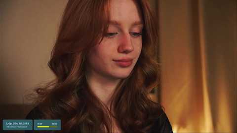 Media: Video of a young woman with fair skin and long, wavy red hair, wearing a black top, smiling gently. Background features a blurred, warm-toned curtain.