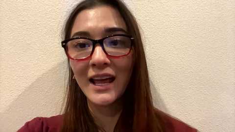 Media: Video of a young woman with long brown hair, wearing red-framed glasses, maroon top, and white headband, smiling, against a textured beige wall.